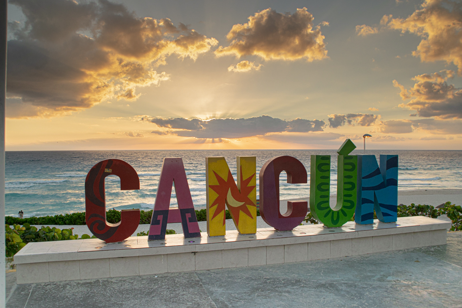 A large, colorful "CANCÚN" sign stands on a stone platform, with the turquoise ocean and a beautiful sunset in the background. The sky is filled with golden and orange clouds as the sun sets over the water