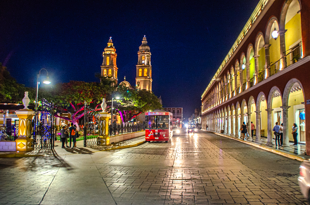  A vibrant night scene in Campeche with a brightly lit cathedral, a cobblestone street, and a tourist trolley