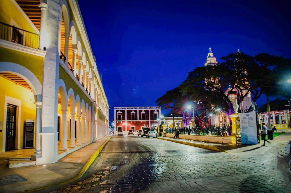 A cobblestone street in Campeche at night, with illuminated colonial buildings and a church spire against a deep blue sky