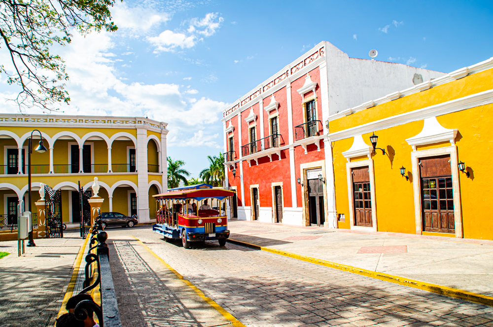A red, white, and blue open-air trolley filled with tourists travels along a street in the historic city of Campeche, with brightly painted buildings on either side under a blue sky