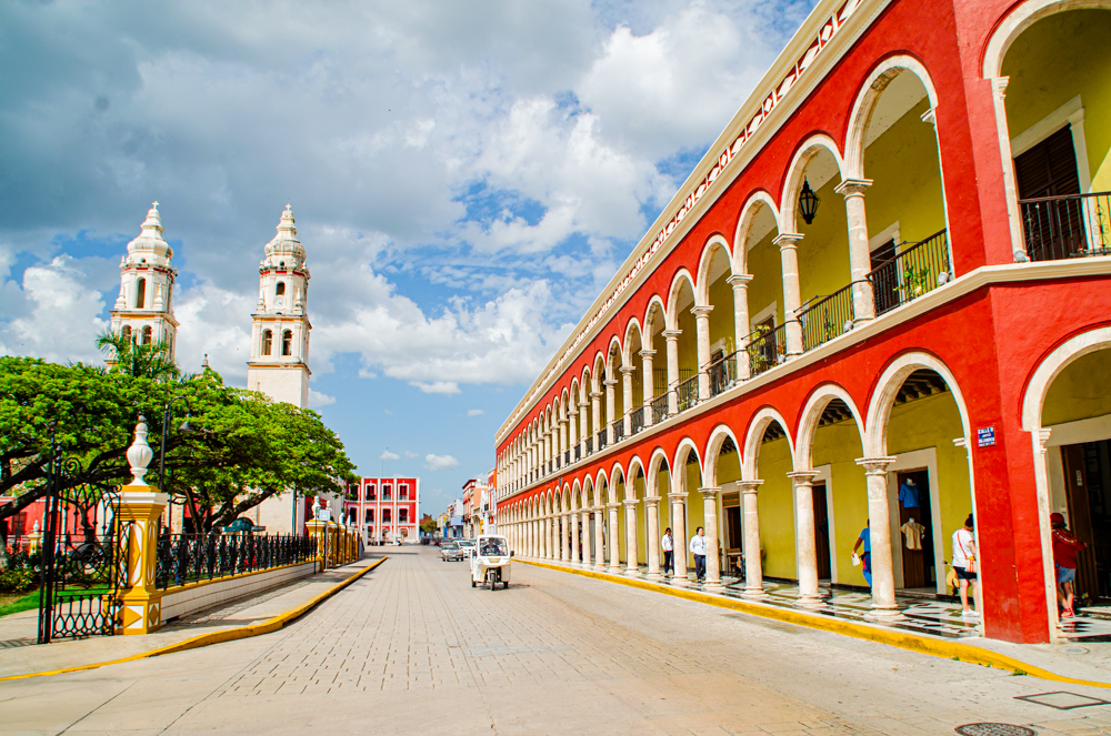 A vibrant street scene in Campeche, Mexico, showing the City Hall, a row of red and yellow archways on the right and a white stone cathedral with two prominent bell towers on the left. 