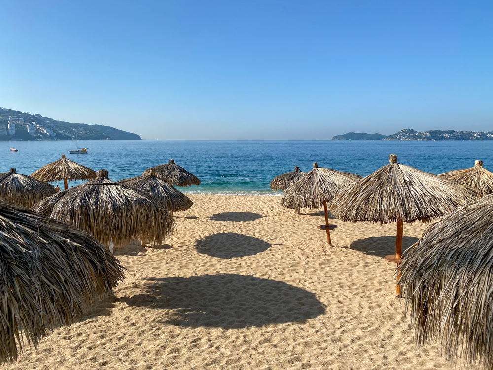 Palapa umbrellas on a sandy beach in Acapulco with the ocean and a blue sky in the background