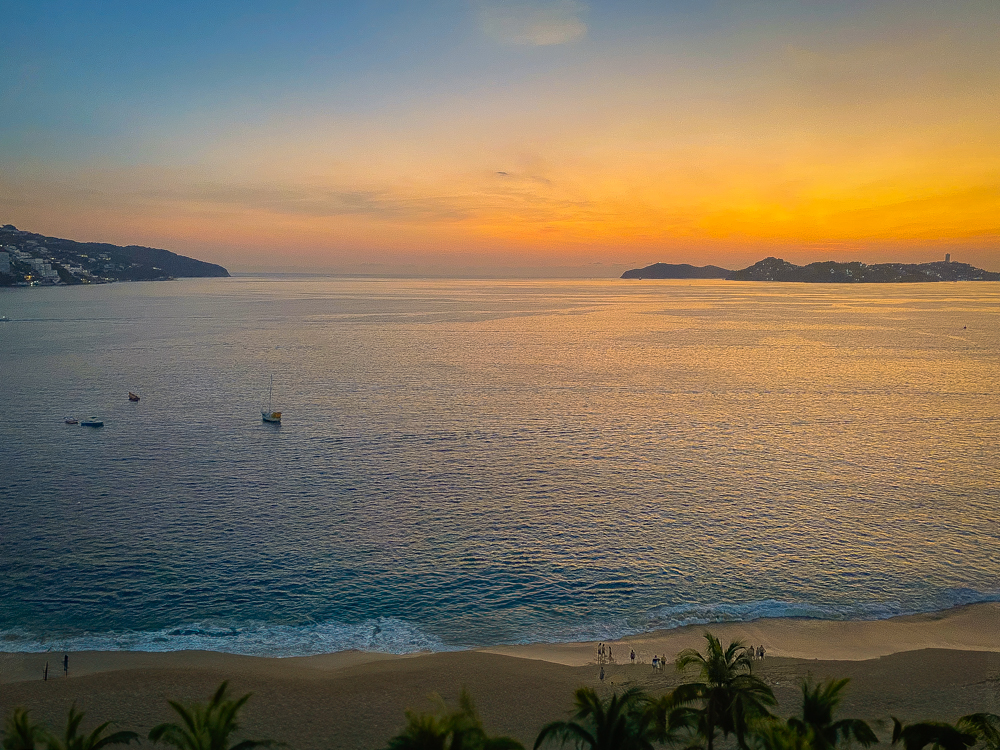 A vibrant sunset over the tranquil Acapulco Bay, with the ocean reflecting the orange and blue sky