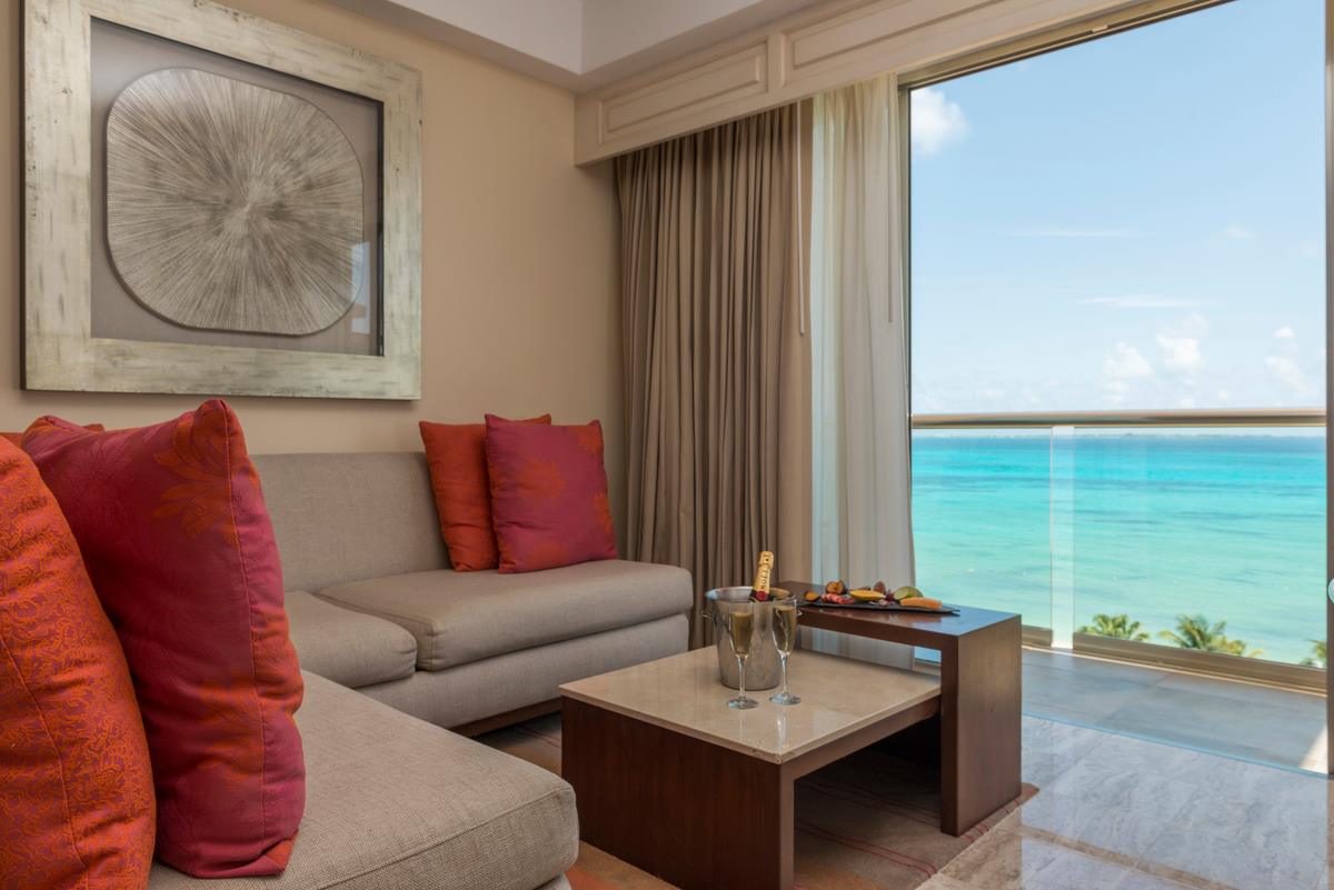 A luxurious and elegant living area of a hotel suite with a beige sofa, vibrant red pillows, and a modern coffee table. A bucket of champagne with two glasses and a small platter of fruit are on a side table. The room opens to a private balcony with a stunning, unobstructed view of the turquoise Caribbean Sea. This image highlights the high-end accommodations and hospitality services, making it an excellent choice for MICE industry executive retreats, corporate incentive trips, and luxury group travel.