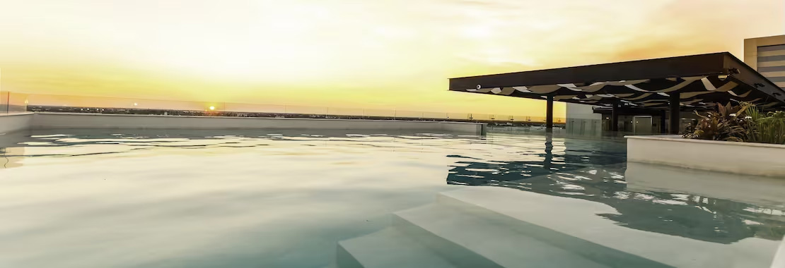 A panoramic rooftop swimming pool at sunset, featuring clear blue water and an open-air shaded lounge area. This luxurious setting is an ideal venue for incentive travel groups, corporate networking receptions, and MICE relaxation. Keywords: Rooftop venue, incentive program, corporate events, sunset reception, networking space, hotel pool, Merida.