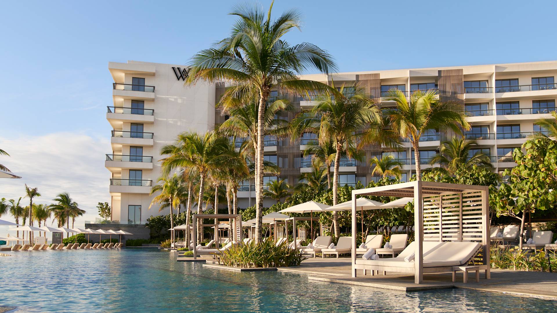 Luxurious poolside cabanas and lounge chairs at The Waldorf Astoria Riviera Maya in Cancun, Mexico. The resort offers a premier meeting and event venue for corporate groups, incentives, and conferences (MICE). The modern hotel building, framed by tall palm trees, overlooks the expansive pool area, ideal for networking receptions and exclusive events