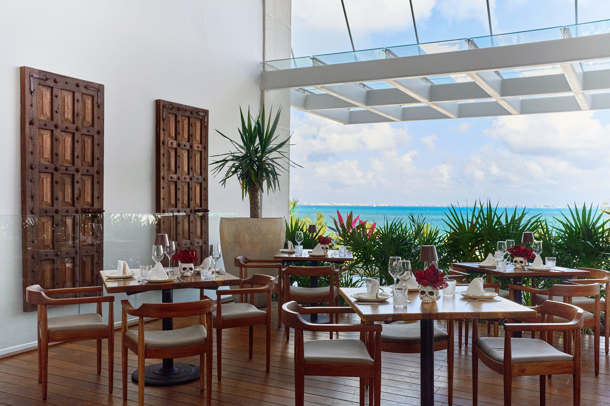 Outdoor terrace dining area at SLS Playa Mujeres with a stunning view of the Caribbean Sea and lush greenery. The sophisticated event space features dark wood floors, modern wood chairs with beige cushions, square tables set with wine glasses and distinctive red floral centerpieces. The open-air design with a glass roof provides an elegant setting for private group dining, corporate dinner events, and high-end networking receptions within the MICE program