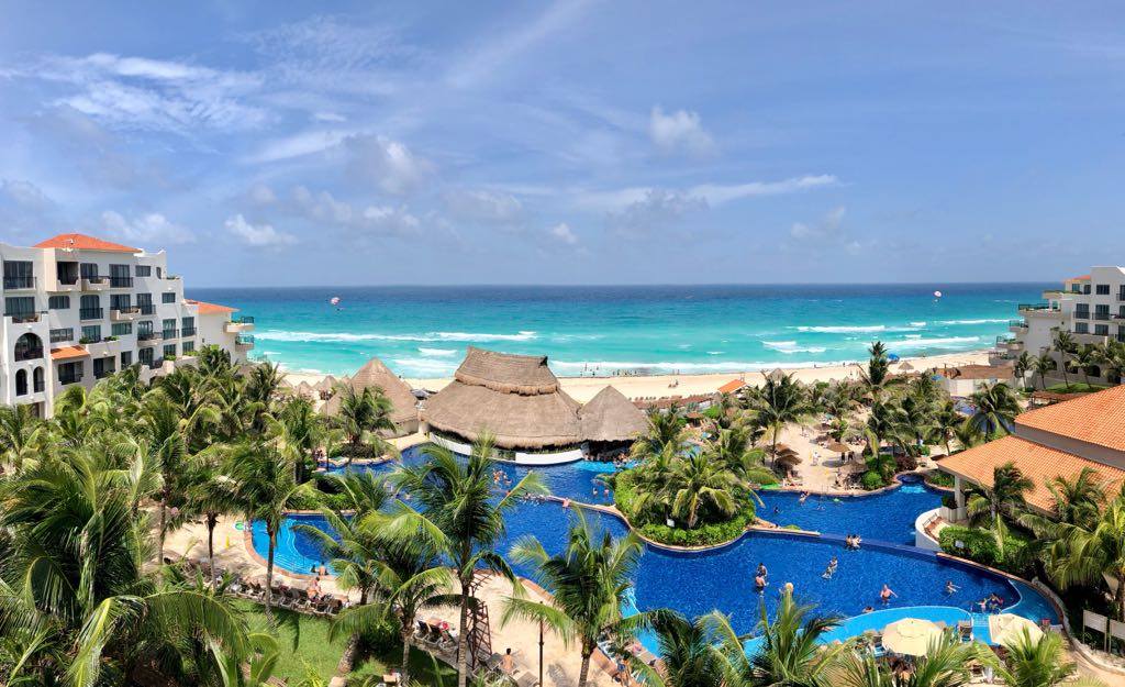 An elevated, panoramic view of the Fiesta Americana Condesa Cancun resort, showcasing a large, multi-level swimming pool with a swim-up bar and guests relaxing. The resort buildings with white facades and palm trees surround the pool area, leading to a pristine white-sand beach and the turquoise Caribbean Sea. This image highlights the breathtaking oceanfront location and leisure amenities, making it an excellent venue for MICE industry events, corporate incentives, and group travel experiences.