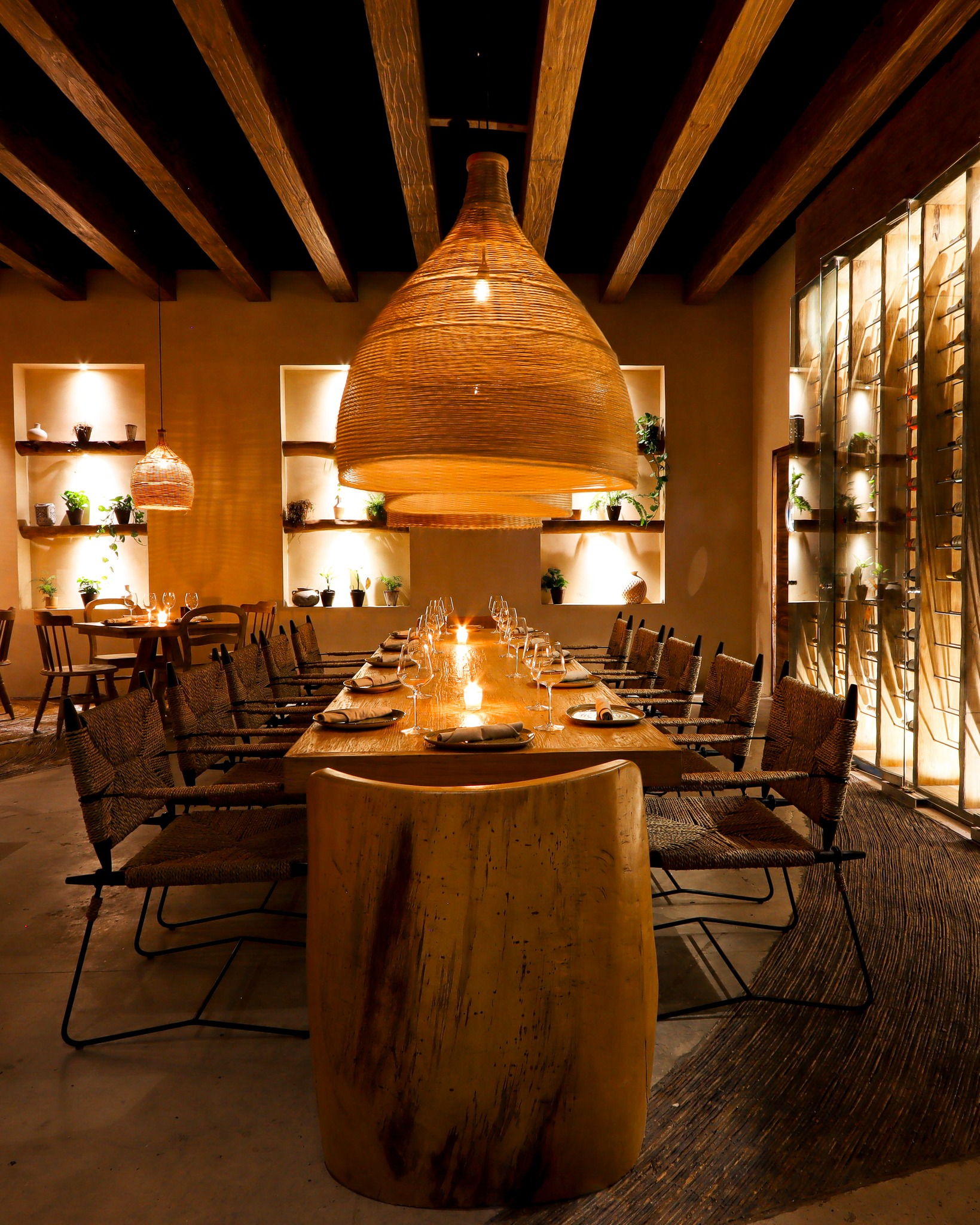 Interior view of an intimate, private dining room set up for a corporate event, featuring a long wooden table with rustic woven chairs and a large, striking organic pendant lamp. The space is elegantly styled with recessed shelving and a backlit wine wall. This premium venue is perfect for small MICE groups, confidential executive meetings, incentive program dinners, and high-level corporate hospitality in Cancún