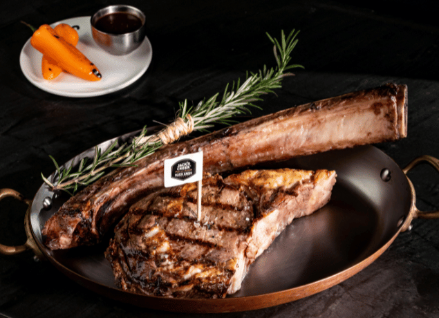 Close-up of a massive, perfectly grilled Tomahawk steak served on a copper platter, garnished with rosemary and featuring a quality brand flag. This image highlights the gourmet catering and premium menu options available for MICE industry events, including luxurious executive dinners, corporate hospitality functions, and incentive travel group dining experiences in Cancún