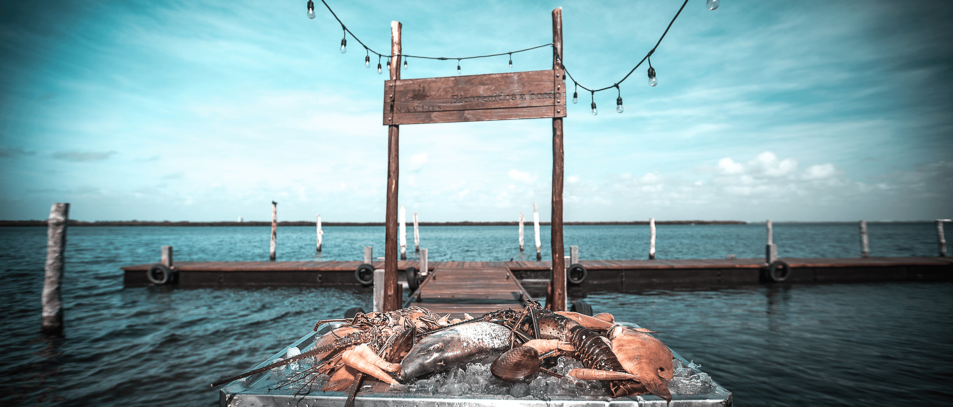 A display of fresh, raw seafood, including lobster and fish, presented on a bed of ice on a metal cart at the edge of a wooden pier or dock. In the background, a sign hangs from a wooden archway with string lights over the calm lagoon waters. This image promotes the premium catering and unique local culinary experiences offered for MICE industry gala dinners, incentive travel events, and executive receptions in Cancún