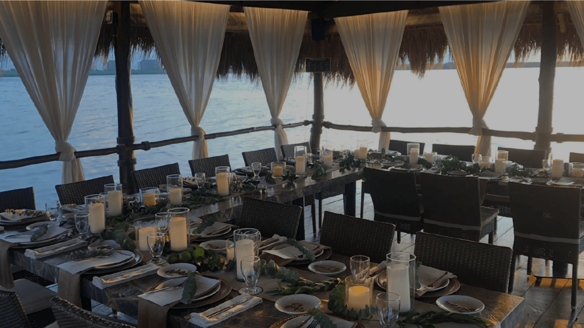 Close-up of an elegantly set dining table in a private, over-the-water palapa structure at sunset, featuring white flowing curtains, candlelight, and green foliage centerpieces. The formal setup is ideal for exclusive MICE industry events, including incentive program farewell dinners, confidential executive meetings, and sophisticated corporate hospitality functions for small groups in Cancún