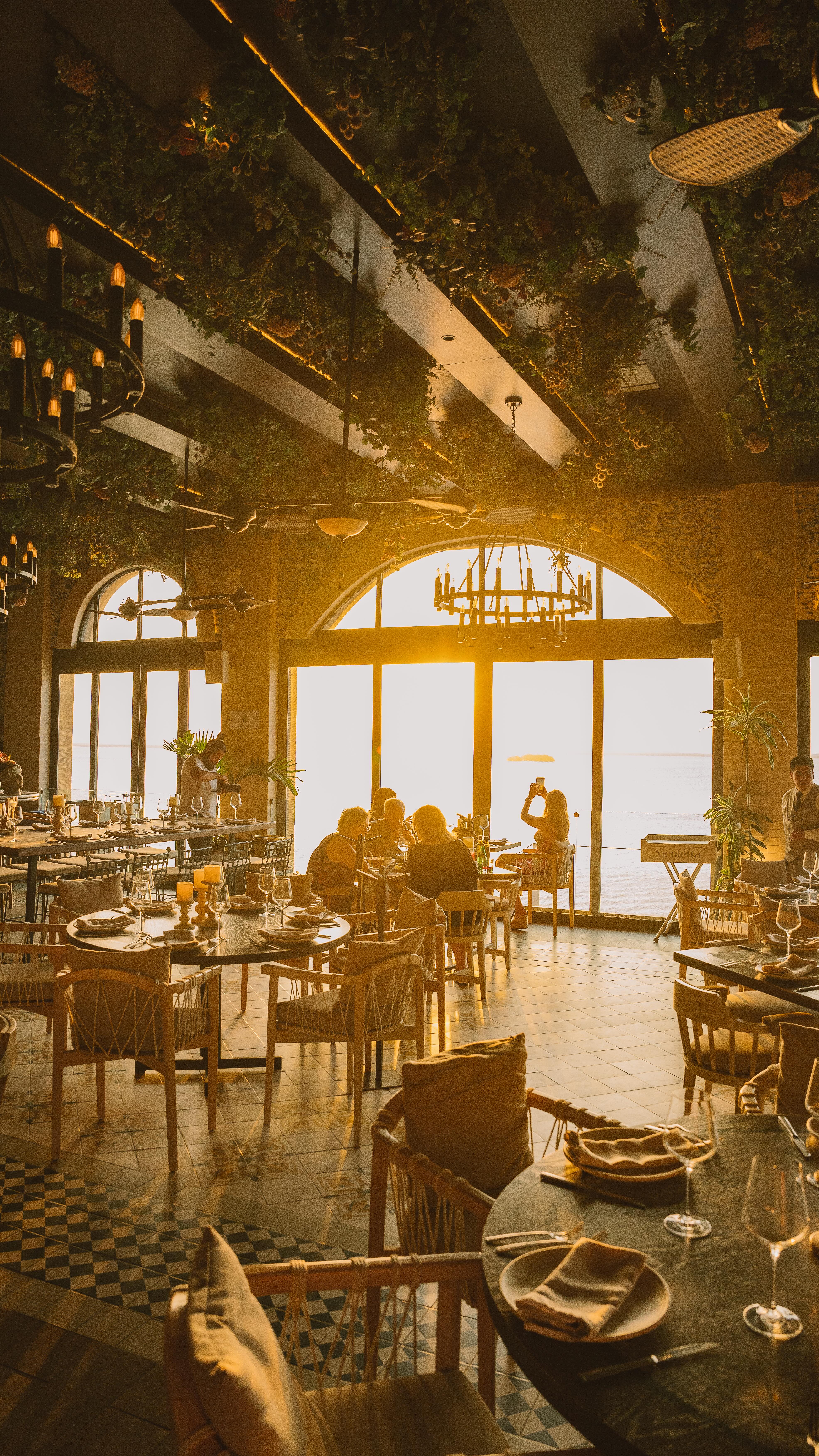 Interior view of Nicoletta Restaurant in Cancún, bathed in the warm, golden light of sunset streaming through large arched windows overlooking the water. The room features elegant tables set for dinner, with greenery hanging from the ceiling and rustic chandeliers. This captivating atmosphere is perfect for MICE groups, creating a memorable backdrop for incentive program welcome receptions, executive dinners, and high-end corporate hospitality.