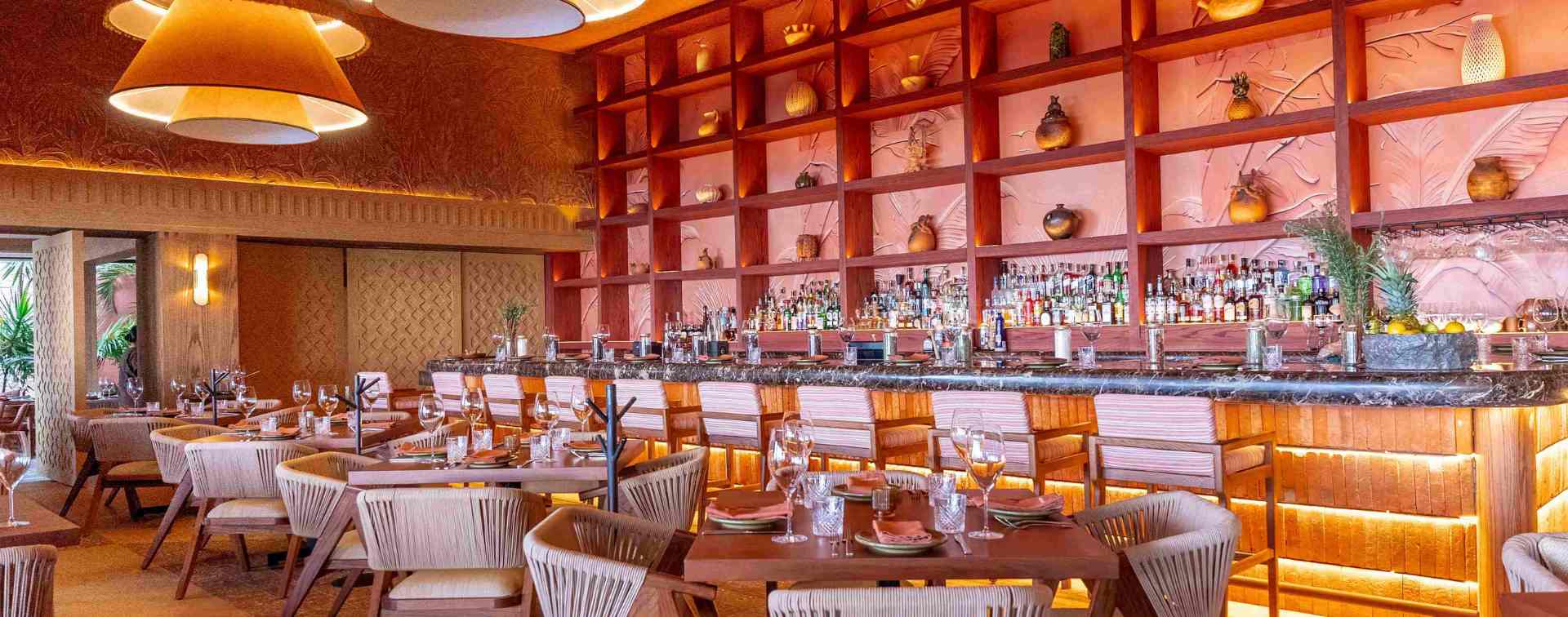 Interior view of Porfirio's Restaurant featuring a long, well-stocked bar with warm lighting, set against a stunning wall of open shelves displaying decorative pottery. The foreground shows tables set for dining or cocktails. This upscale space is ideal for MICE industry networking receptions, executive cocktail hours, and corporate welcome mixers, highlighting a vibrant and luxurious atmosphere for corporate hospitality in Cancún.