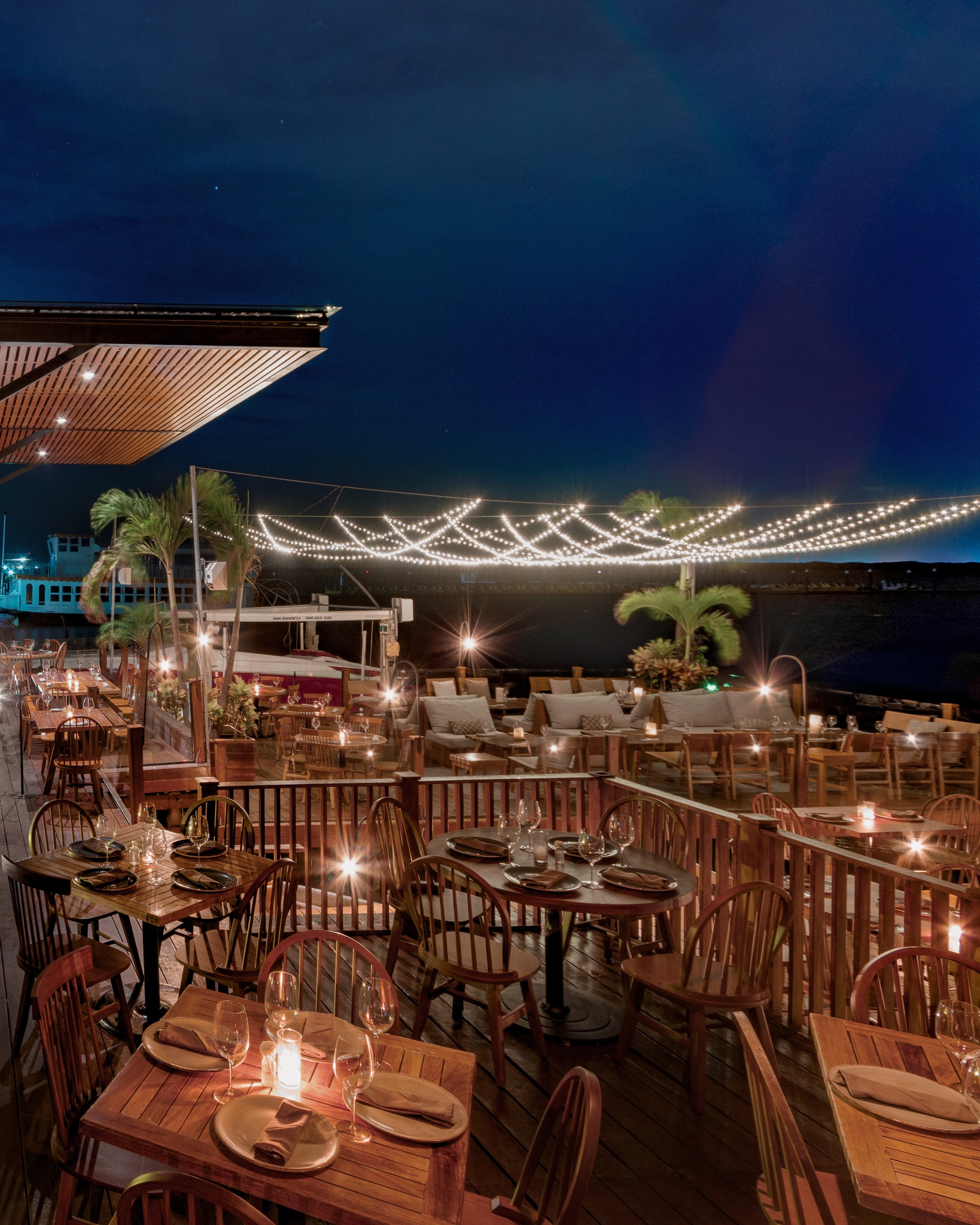 Night view of a multi-level outdoor dining terrace at RosaNegra Restaurant, overlooking the dark water, dramatically lit by string lights hanging overhead. The spacious wooden deck features numerous tables set with candles, demonstrating high capacity for large MICE industry functions, corporate gala dinners, and incentive travel closing banquets in a festive, open-air Cancún setting.