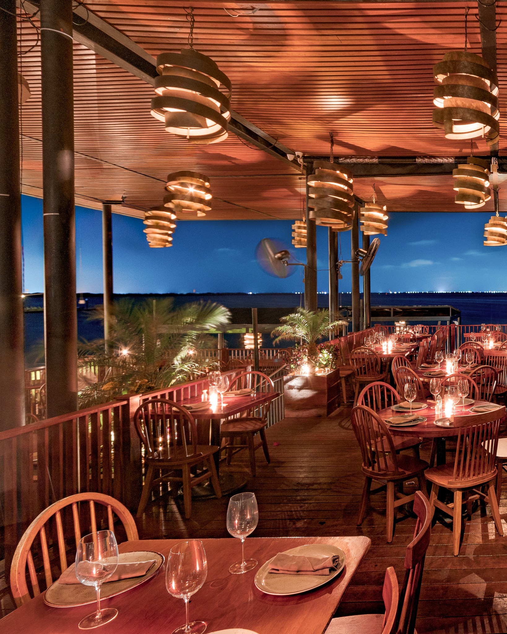 Exterior night view of a luxurious, open-air terrace at RosaNegra Restaurant overlooking the water. The space is warmly lit with unique coiled pendant lamps, candlelight, and dark wood furniture. The intimate table settings are ideal for MICE industry groups hosting exclusive corporate dinners, incentive travel receptions, or high-end networking events in a sophisticated Cancún waterfront atmosphere.