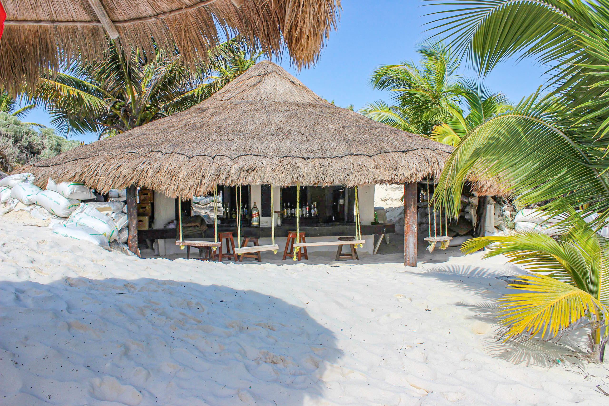 Quaint thatched-roof beach bar hut in Tulum, Mexico, surrounded by white sand, palm fronds, and stacked bags, featuring wooden stools, hanging bar swings, and bottled drinks— a cozy venue for MICE networking mixers, incentive travel cocktail hours, and informal conference breaks in an authentic Caribbean setting