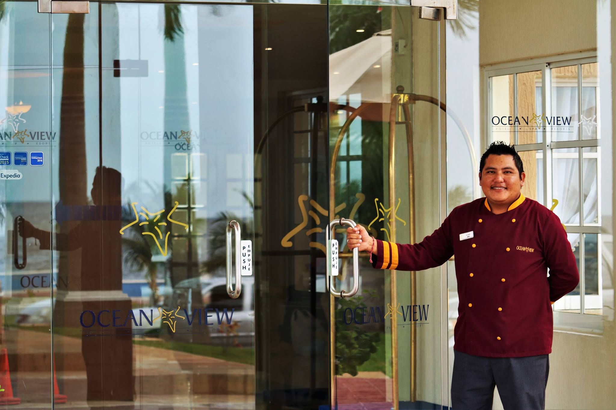 Bell staff member in uniform smiling and holding open the clear glass doors of the Ocean View Hotel's main entrance in Campeche. The doors feature the hotel's logo, and reflections show the outside area and pool. This image emphasizes the friendly, personalized service and smooth logistics provided for MICE attendees, corporate groups, and incentive travelers upon arrival in Mexico