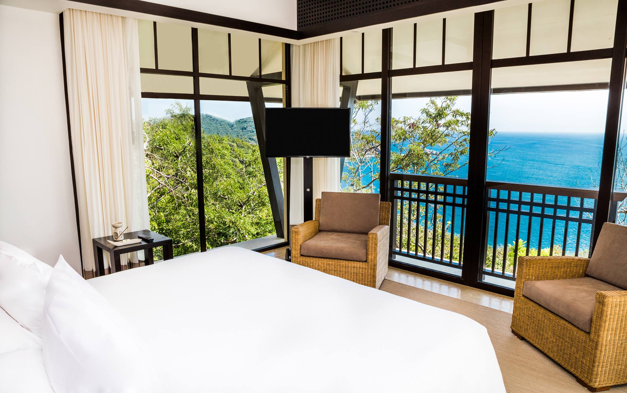 A luxurious king bedroom at the Banyan Tree Cabo Marques, featuring a large white bed, rattan chairs, and floor-to-ceiling windows with stunning views of the ocean and lush tropical hills. This premium accommodation provides a high-end, relaxing escape for incentive travel winners or VIP MICE delegates, emphasizing luxury and comfort after a successful corporate event.