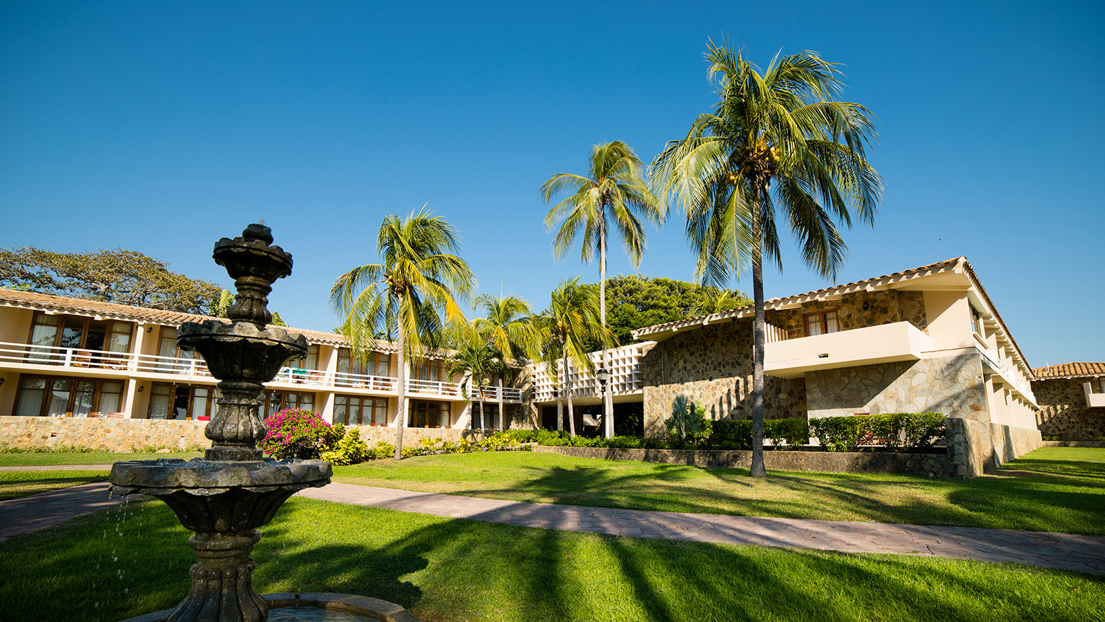 A low-angle view of the central courtyard and grounds of The Pierre Mundo Imperial resort in Acapulco, featuring lush green lawns, tall palm trees, and low-rise stone and stucco hotel buildings. A classic decorative stone fountain is visible in the foreground. This tranquil and beautiful setting is ideal for hosting incentive travel groups, offering ample space for outdoor team building activities, and providing a relaxing atmosphere for MICE delegates.