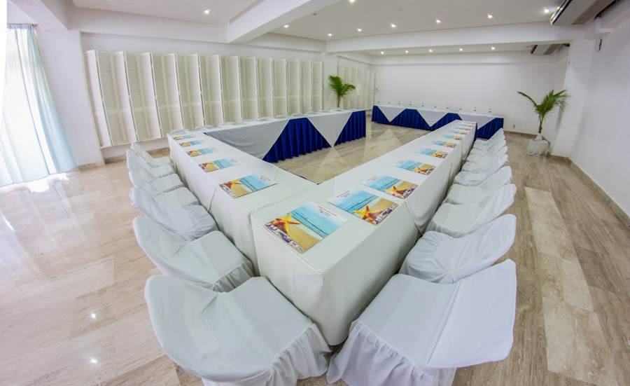 A bright, modern meeting room at the Playa Suites Hotel in Acapulco, configured in a U-Shape style for group interaction. The room features a white color palette, marble-like flooring, and white-skirted chairs and tables, providing an elegant and professional environment. This setup is ideal for focused board meetings, interactive training sessions, and high-level MICE discussions.
