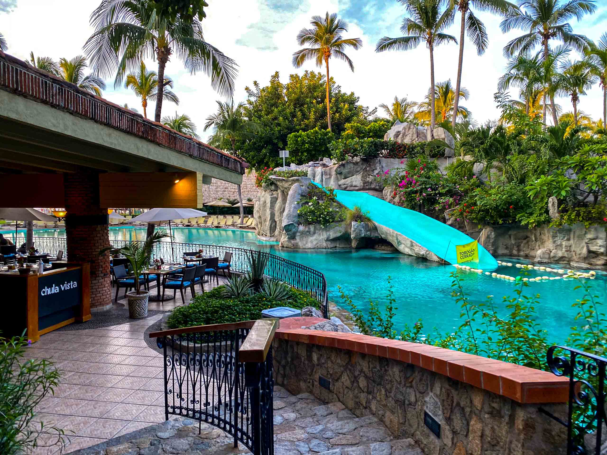 A lush, tropical resort pool area in Acapulco featuring a large, blue freeform pool with a decorative rock waterfall and a bright blue waterslide. The pool deck includes a stone staircase and a covered outdoor bar/dining area, creating an inviting setting. This fun and vibrant environment is ideal for incentive travel groups, providing excellent leisure and delegate downtime activities at a premier corporate retreat destination.