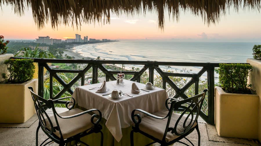 A close-up view of an elegantly set table for two on a private balcony at the Quinta Real Acapulco, overlooking the sprawling bay and beach at sunset. The table setting, featuring white linen and glassware, is framed by a rustic railing and a palapa-style thatched roof. This image highlights the premium, gourmet dining and exclusive corporate hospitality experiences available for incentive travel groups and VIP MICE delegates.