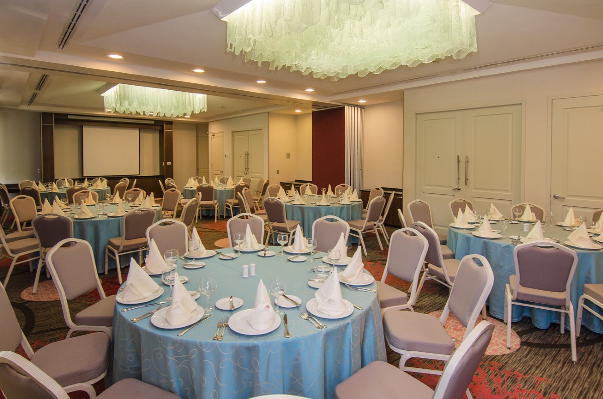 An interior view of a versatile meeting room at the Hilton Garden Inn, set up for a banquet function in a round table configuration. The room, ideal for corporate dinners, gala events, or conference lunches, features elegant blue tablecloths, contemporary chairs, and a retractable screen for presentations. This shows a flexible event space suitable for various MICE industry needs.