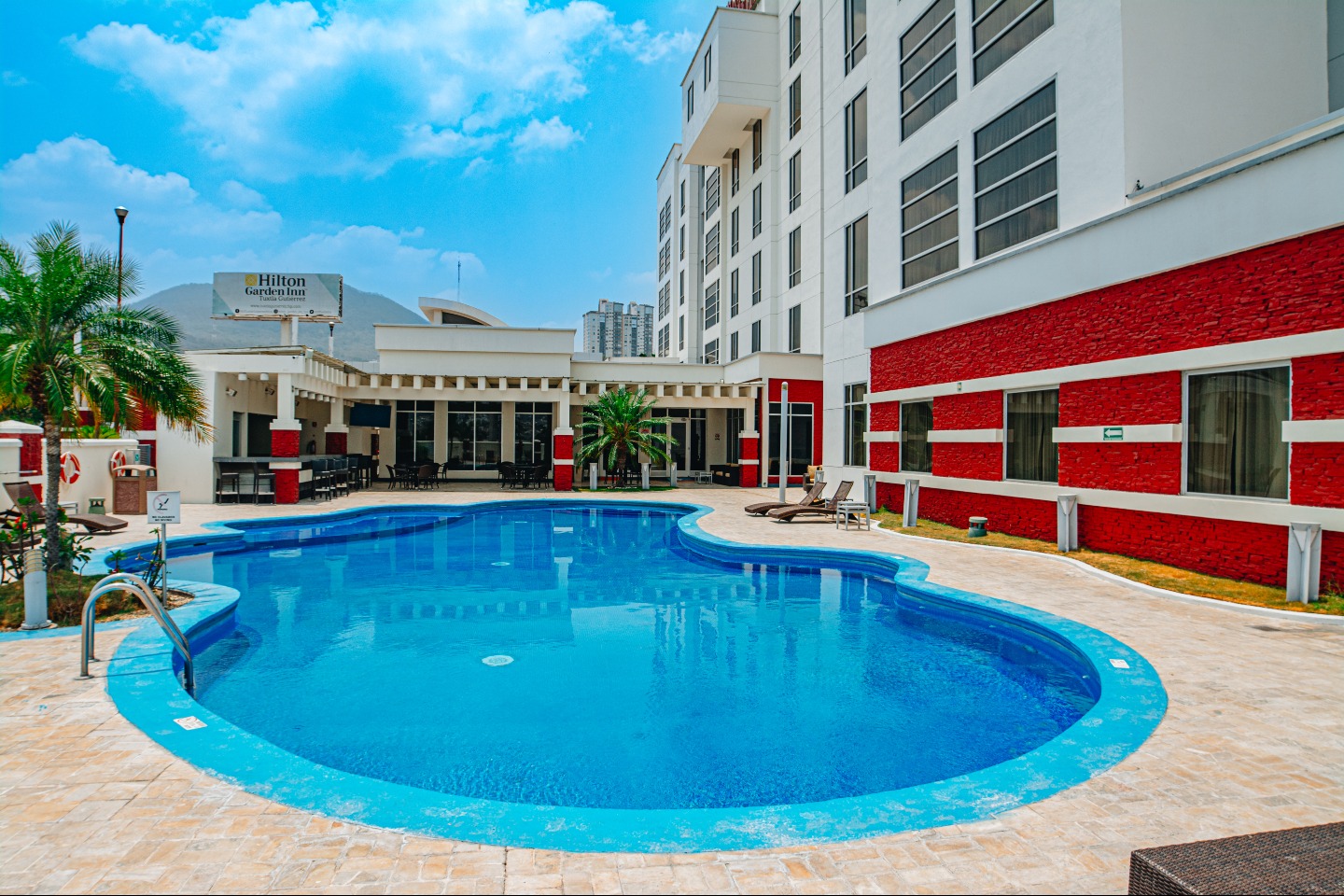 The outdoor swimming pool area of the Hilton Garden Inn hotel, featuring a bright blue pool surrounded by sun loungers and tropical foliage. This attractive leisure space offers essential delegate downtime and is perfect for unwinding after meetings or as a relaxed setting for incentive group activities. The modern hotel building, with red accent brickwork, provides a comfortable and comprehensive environment for MICE attendees.