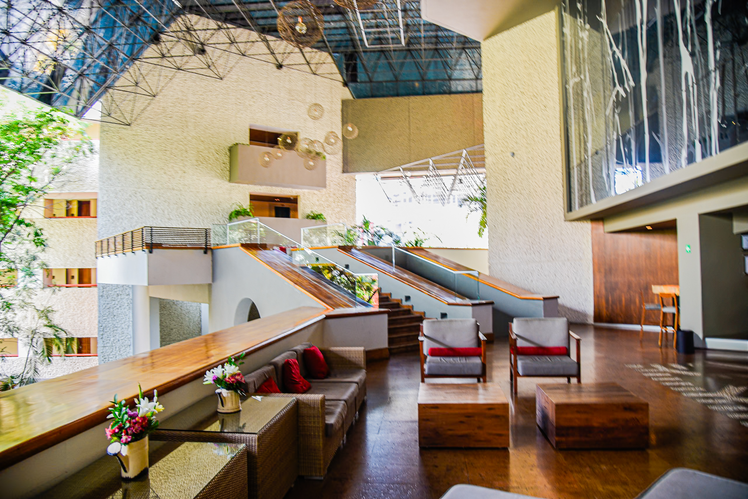 The bright, high-ceilinged interior lobby and common area of the Marriott Tuxtla Gutiérrez. The modern design features natural light from a large glass roof, rich wood floors, comfortable seating areas (ideal for small group meetings and informal networking), and tiered access via stairs and escalators. This spacious area is perfect for attendee registration, large conference breaks, and creating a welcoming atmosphere for MICE delegates.