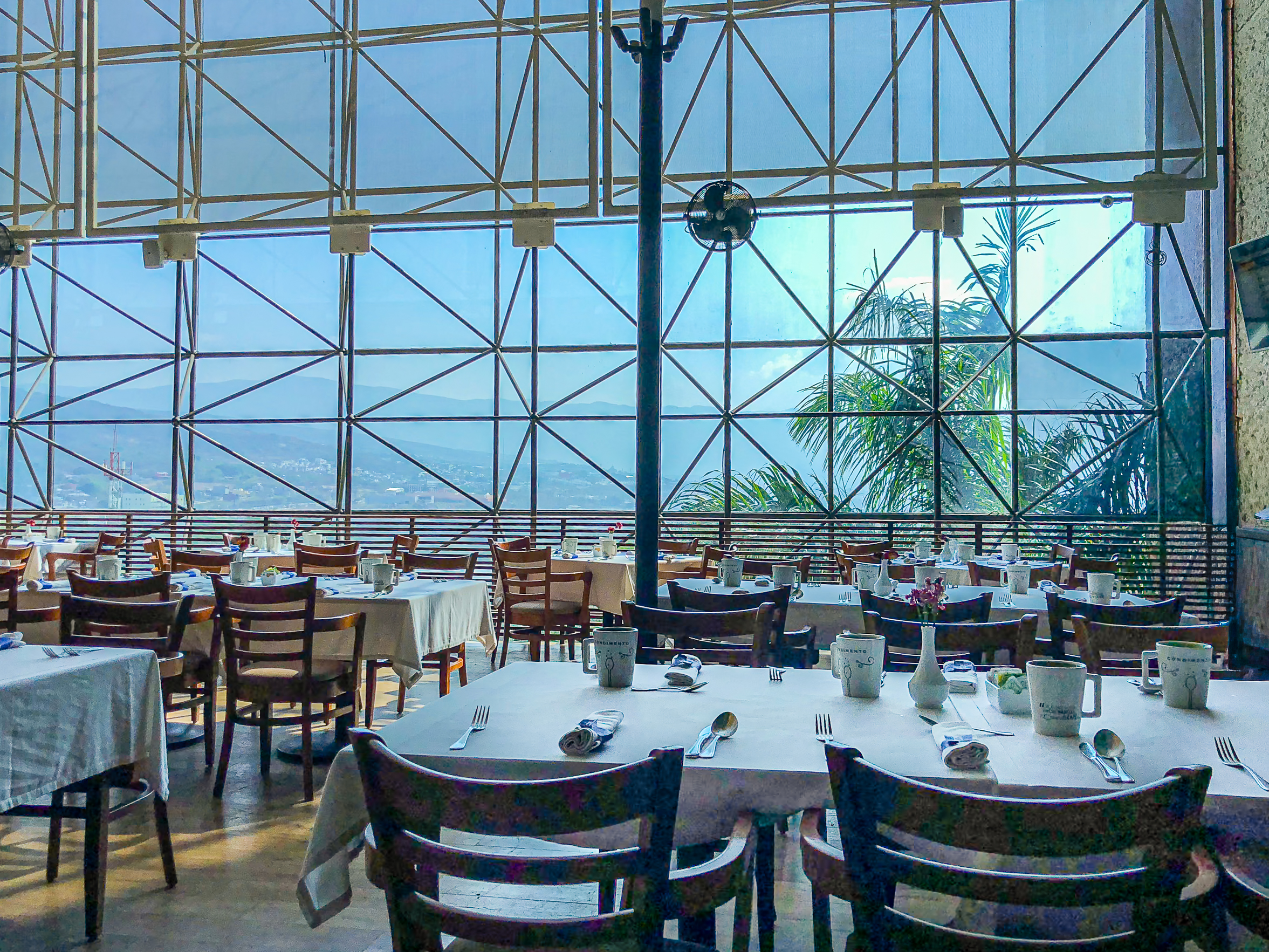 The bright dining area of the Marriott Tuxtla Gutiérrez, set up with multiple tables for a breakfast or group dining function. The room features a dramatic, floor-to-ceiling glass wall with metal framing that offers a panoramic view of the mountains and city skyline. This distinctive setting is perfect for memorable delegate meals, conference lunches, or exclusive incentive group dining experiences.