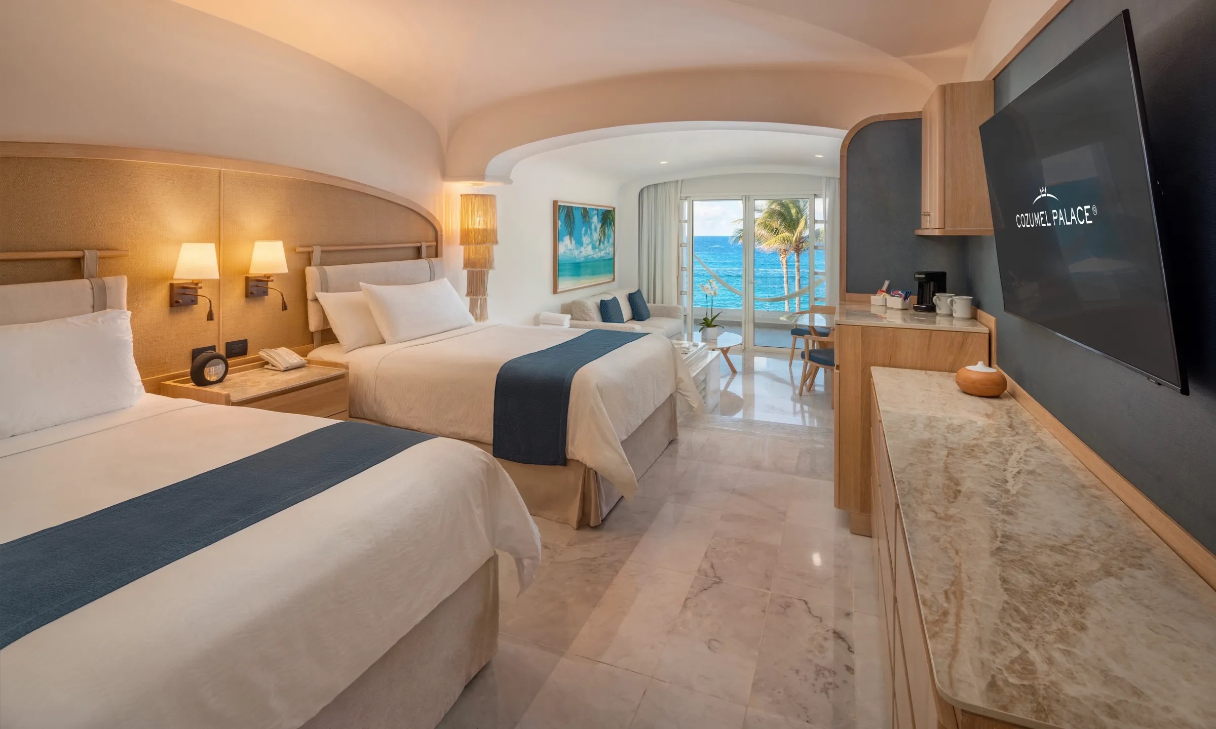A bright, luxurious guest room at Cozumel Palace, featuring two double beds with marble flooring, modern wood cabinetry, and a seating area in the background that opens directly to an ocean view. The room is spacious and designed for comfort, making it an excellent choice for MICE delegates or incentive group attendees sharing accommodation while providing easy access to resort amenities.