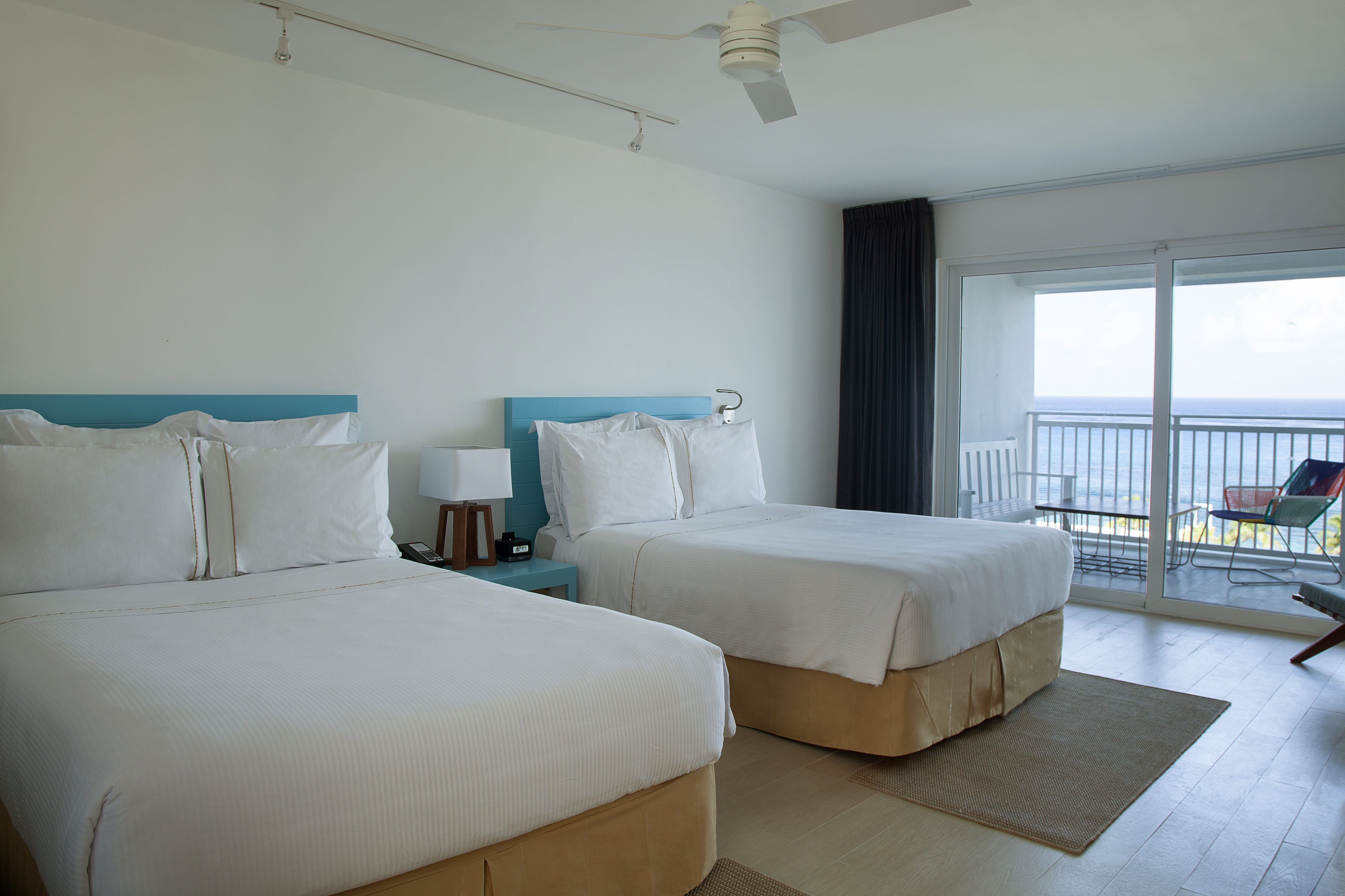 A bright, minimalist guest room at the Fiesta Americana Cozumel featuring two double beds with white linens, light wood floors, and modern tropical decor. A large sliding glass door opens to a private balcony with an ocean view. The setup is ideal for MICE delegates sharing accommodation, providing comfortable and visually appealing lodging for both incentive travel and corporate conference attendees.