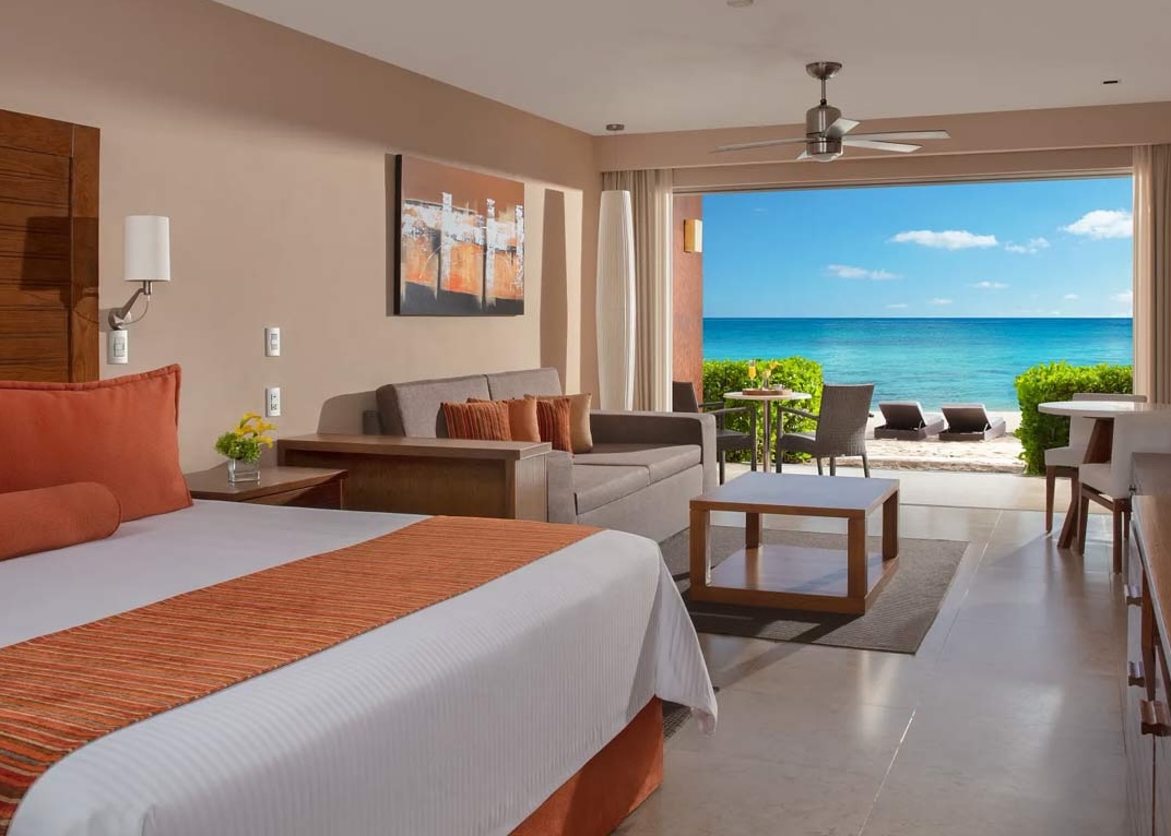 A luxurious, ground-floor beachfront suite at Secrets Aura Cozumel, featuring a king bed and a comfortable sitting area with a couch and coffee table. Large sliding doors open directly onto a private patio with a dining set, leading immediately to the white sand beach and the turquoise ocean. This premium, all-inclusive accommodation is the perfect incentive travel reward, offering direct access and a high-end experience for VIP MICE delegates.