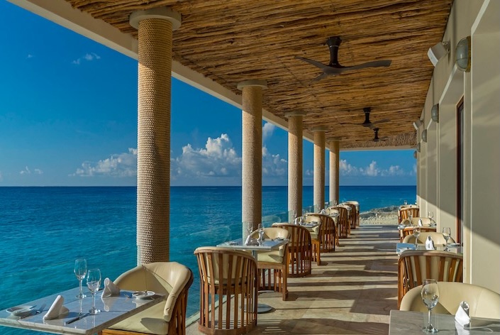 A stunning view of an open-air, covered restaurant deck at The Westin Cozumel, featuring rows of elegantly set tables with wicker chairs and views of the vast, clear blue Caribbean Sea. The ceiling is made of woven straw, supported by rough-textured columns. This luxurious, oceanfront setting is perfect for high-end corporate hospitality, incentive group dining, and memorable MICE networking receptions right on the water.