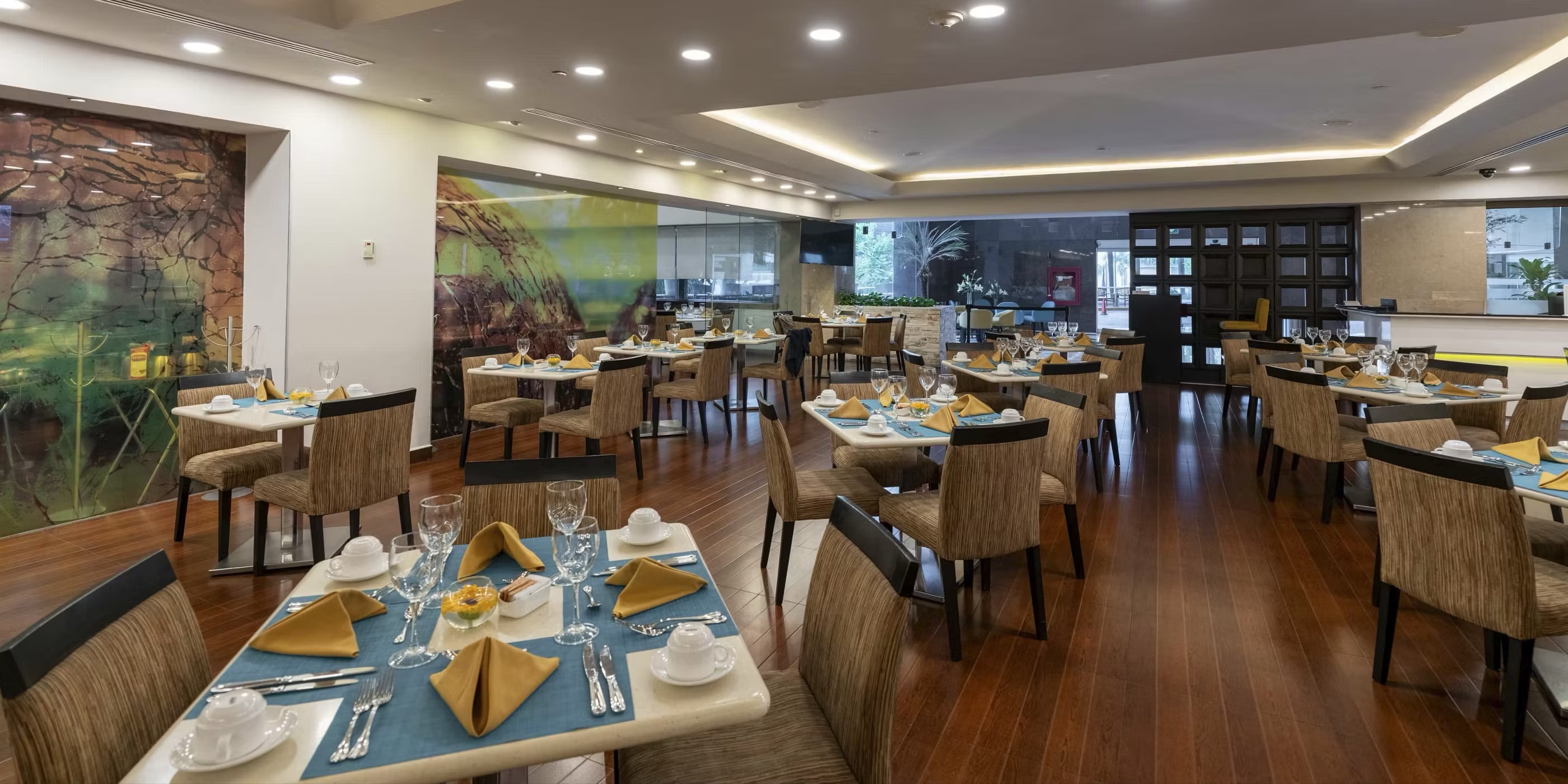 A modern, well-lit restaurant at the Barceló Guadalajara, featuring tables set with blue placements and elegant tableware, ready for group dining. The space includes comfortable chairs and warm wood flooring, offering a professional yet inviting atmosphere for conference meals, business breakfasts, and MICE delegate dining experiences. The contemporary art on the walls adds a touch of local flair to the event venue.