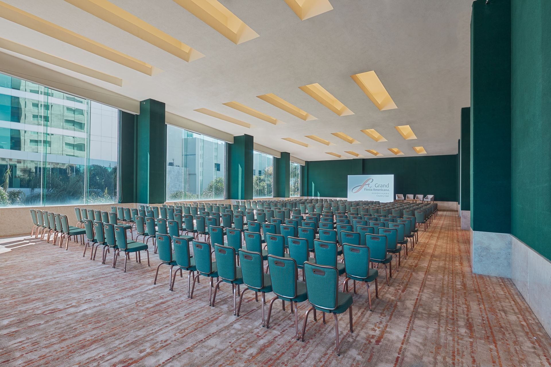 A large, professional meeting room at the Grand Fiesta Americana Guadalajara set up in a theater style with rows of teal chairs, ideal for a conference, seminar, or general session. The room features large windows for natural light (with city views) and a large screen for presentations, showcasing a versatile and spacious event venue that meets the demands of high-level MICE delegates and conventions.