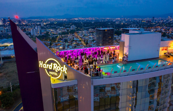 An aerial nighttime view of the spectacular rooftop at the Hard Rock Hotel Guadalajara, featuring a large, illuminated terrace and infinity pool with city views. The area is set up for a dynamic evening reception with a large group of people (incentive travel or networking event attendees). This unique, high-impact space is ideal for high-end MICE events, gala dinners, and memorable corporate celebrations.