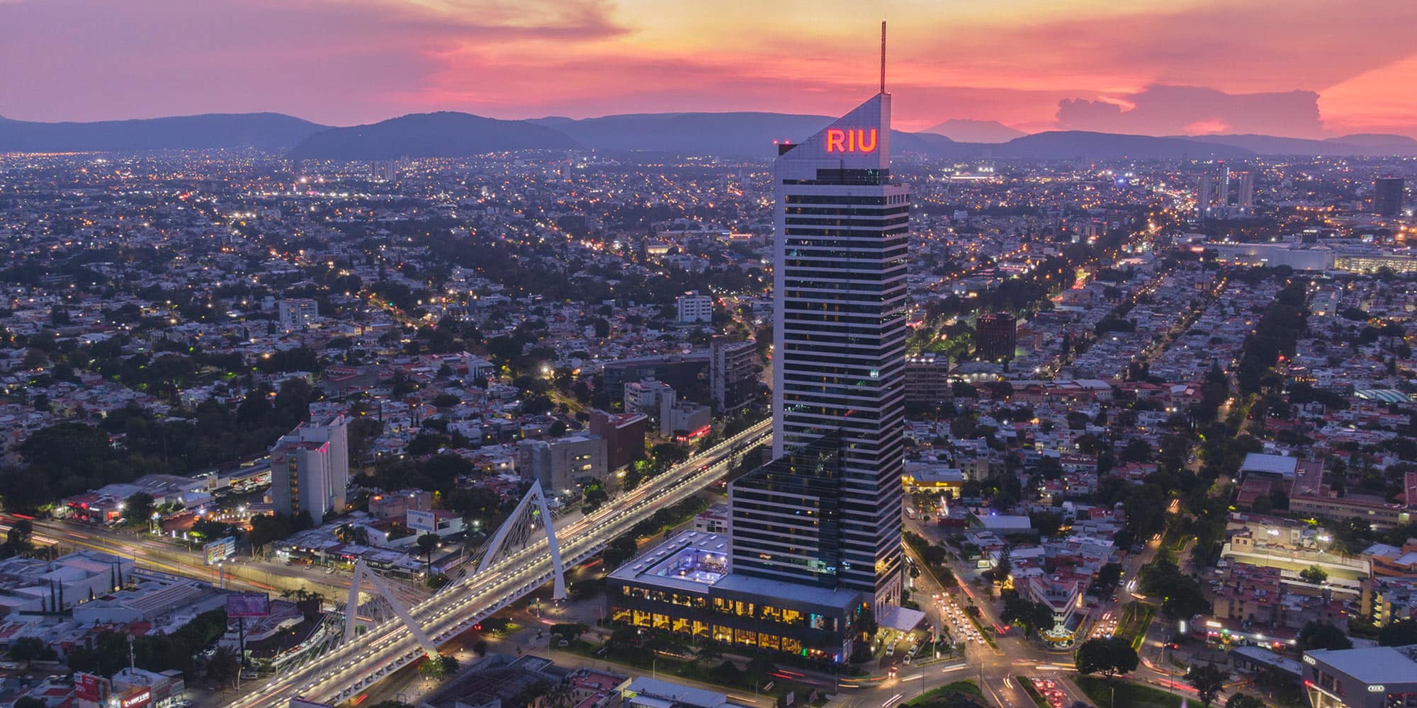 An aerial panoramic shot of the Guadalajara city skyline at dusk, featuring the prominent, illuminated skyscraper of the Hotel Riu Plaza Guadalajara. The high-rise hotel, situated near major urban routes, symbolizes a premier MICE destination offering high-capacity conference facilities, premium accommodations, and excellent city-center access for large conventions and corporate groups.