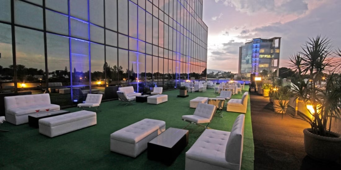 A modern outdoor rooftop lounge area at the Presidente InterContinental Guadalajara at dusk. The space features white modular seating, low tables, and blue accent lighting reflecting on the glass exterior wall, creating a chic ambiance for evening receptions and networking events. This unique outdoor event venue is perfect for premium incentive groups and high-level corporate hospitality with city views.