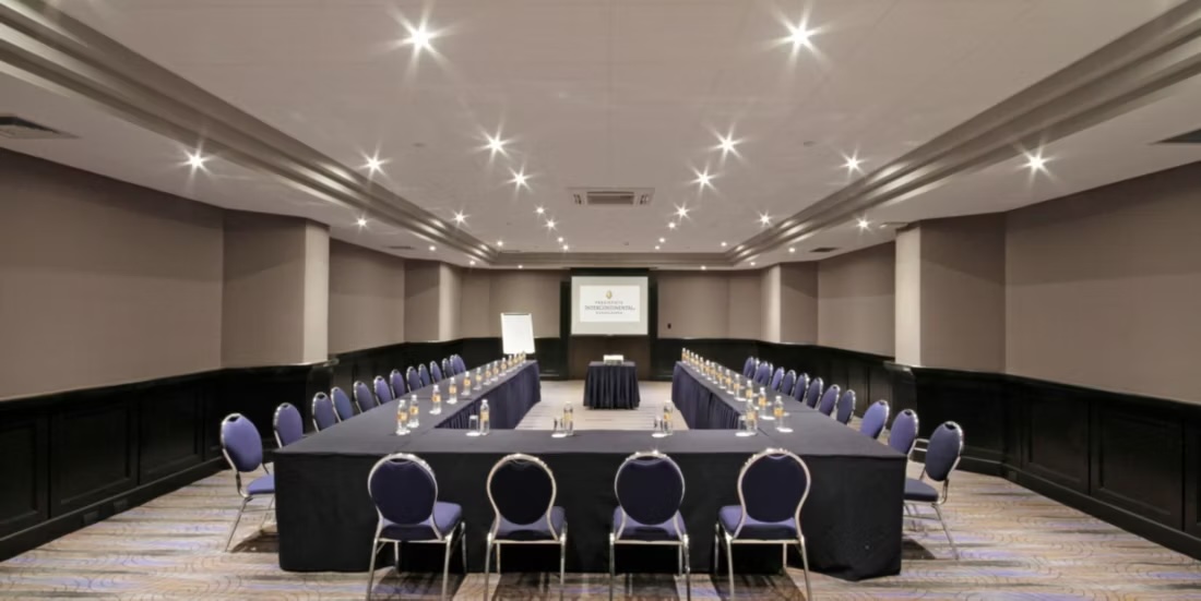 An interior view of a professional, mid-sized meeting room at the Presidente InterContinental Guadalajara, set up in a formal U-Shape configuration with blue chairs and black table linens. This setup is perfect for focused board meetings, training sessions, and high-level corporate discussions. The integrated screen and projector highlight its full readiness as a functional, premium MICE event venue.