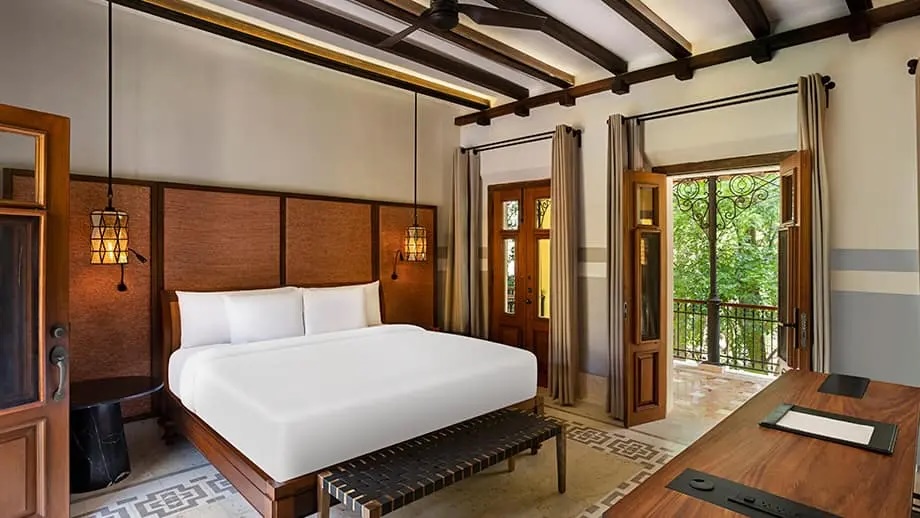 Luxury accommodations for MICE delegates at Hacienda Xcanatun: Interior view of a high-end guest suite featuring a king-size bed with white linens, dark wood furniture, a desk workspace, exposed ceiling beams, and French doors opening to a private balcony, ideal for incentive travel accommodations.