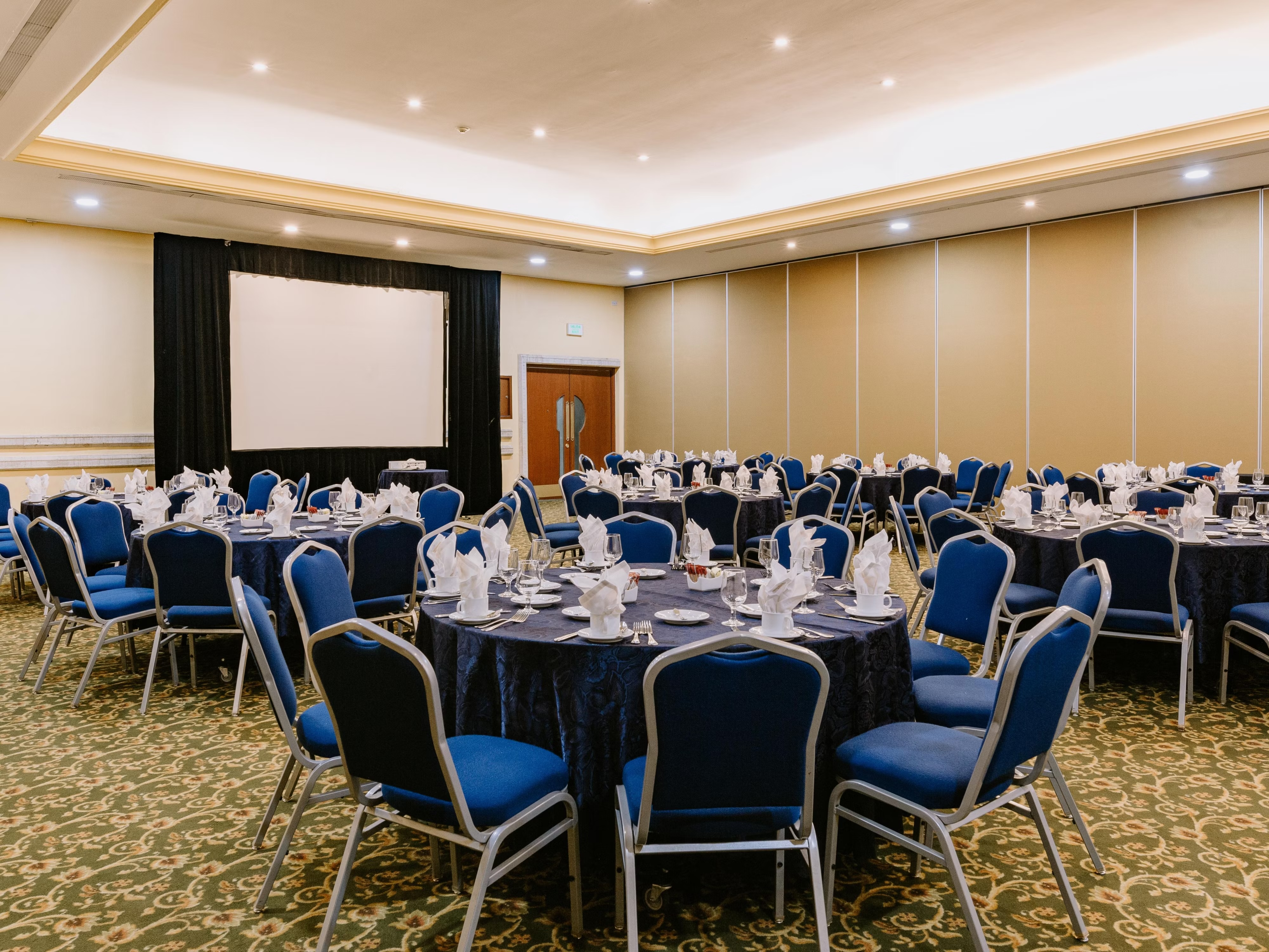 High-capacity convention banquet setup at Holiday Inn Mérida: Interior view of a large hotel meeting room or ballroom set with round tables and blue chairs for a banquet or gala dinner. The space includes a retractable screen for presentations, ideal for large MICE functions and corporate events.