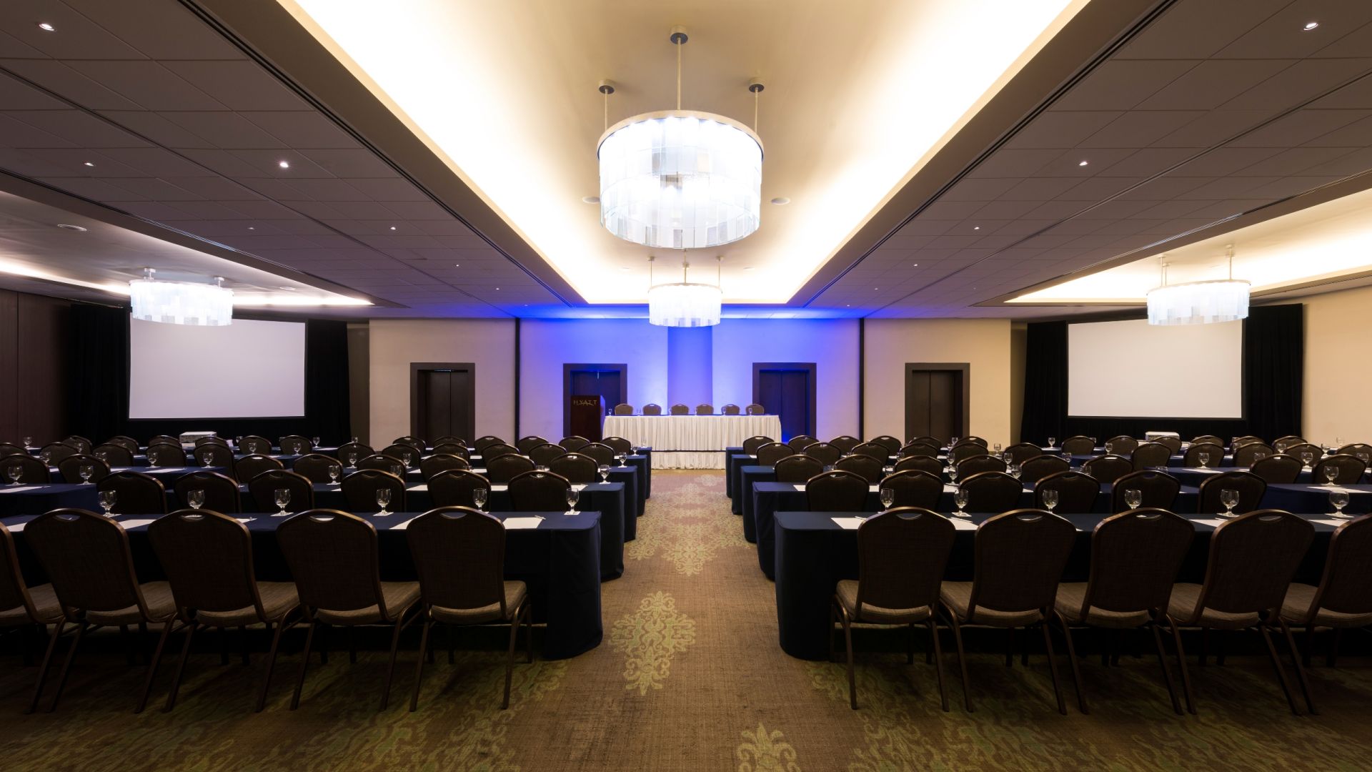 Regency Ballroom setup for a large MICE convention: Wide-angle view of the Hyatt Regency Mérida ballroom configured in a classroom-style arrangement with navy blue tables and chairs, two large projection screens, and modern chandeliers. This setup is ideal for corporate training and plenary convention sessions.