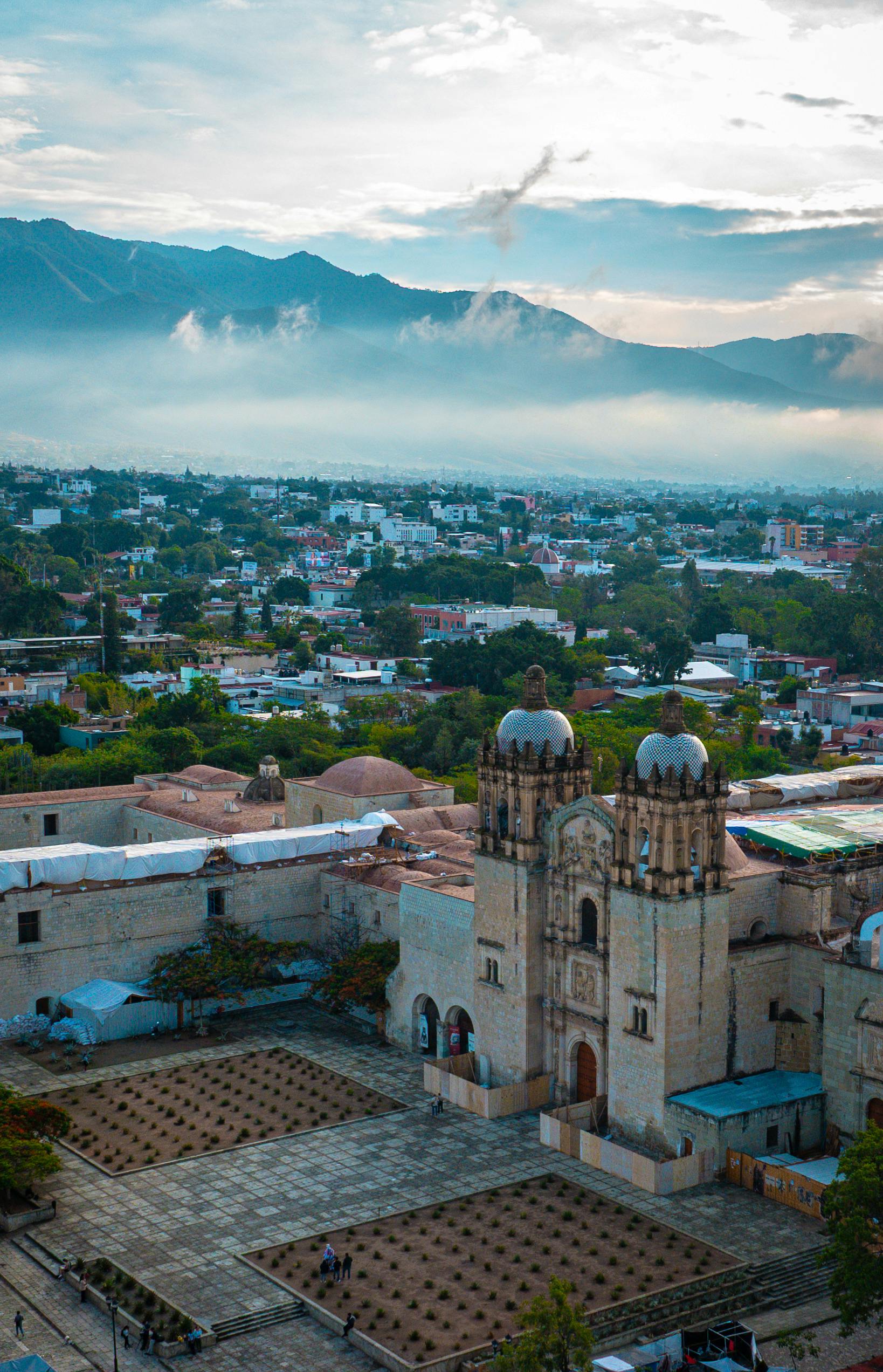 An elevated view of the historic city of Oaxaca, featuring the iconic Templo de Santo Domingo de Guzmán with mountains shrouded in mist in the background. This view highlights the city's colonial charm and its appeal for cultural and historical tours for MICE groups.