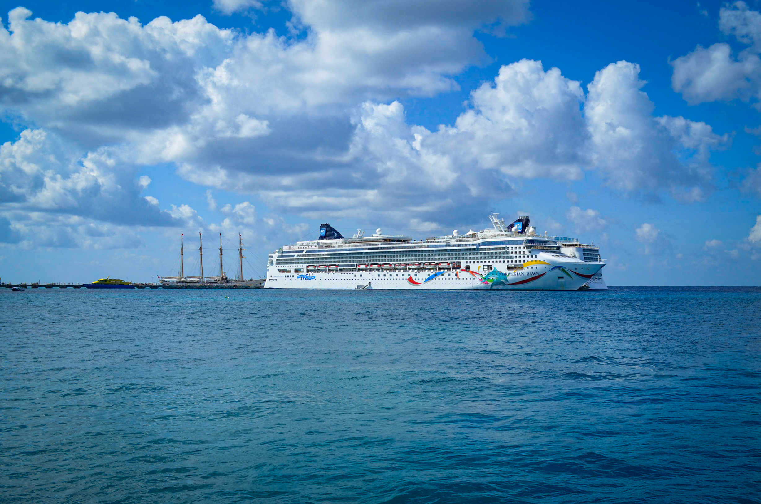 Majestic Norwegian Cruise Ship docked at a vibrant Caribbean port in Cozumel for MICE incentive rewards, corporate conferences, and exhibition events: gleaming white hull with blue funnels, multiple decks with balconies and lifeboats; adjacent historic tall ships with three tall wooden masts and sails furled on a wooden pier extending into turquoise ocean waters; yellow ferry boat nearby; background of deep blue sea merging with azure sky dotted with fluffy white clouds – ultimate floating venue for high-seas meetings, team incentives, and luxury group travel in tropical paradise.