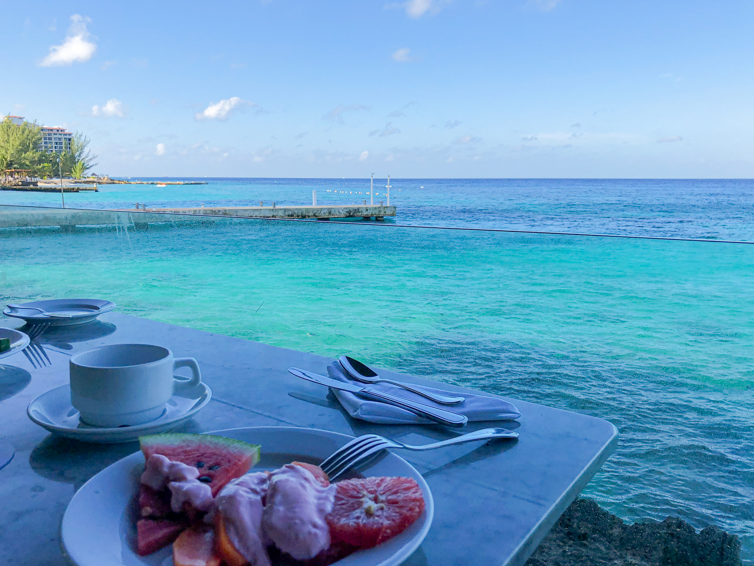 Gourmet tropical breakfast at an ocean front restaurant in Cozumel. Stunning ocean view with turquoise waves against a weathered pier, yellow buoys, deep blue sea, palm-fringed pink hotel building, white sand beach, and clear blue sky – ideal seaside setting for incentive travel, corporate events, exhibitions, and team-building retreats in paradise