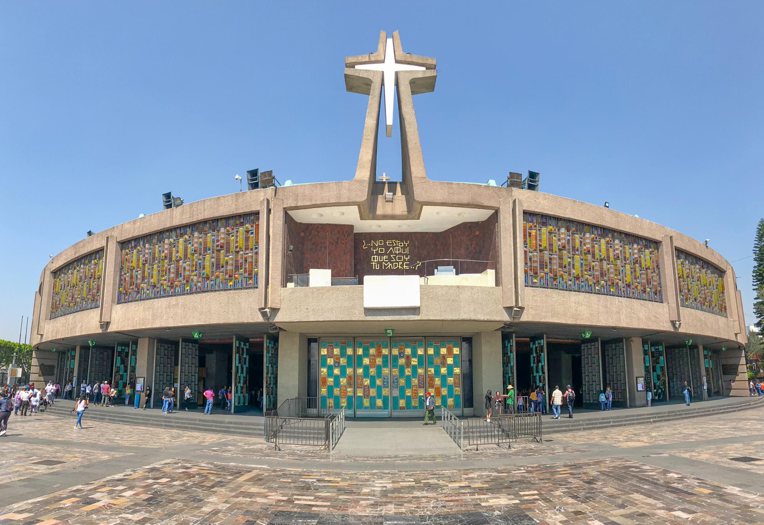 The modern Basilica of Our Lady of Guadalupe in Mexico City, a circular building with a distinctive cross on top. The facade features stained-glass windows and a large entrance. The building is surrounded by a plaza with people walking around. This major religious and cultural landmark is a unique venue to be visites for MICE industry tours and events.