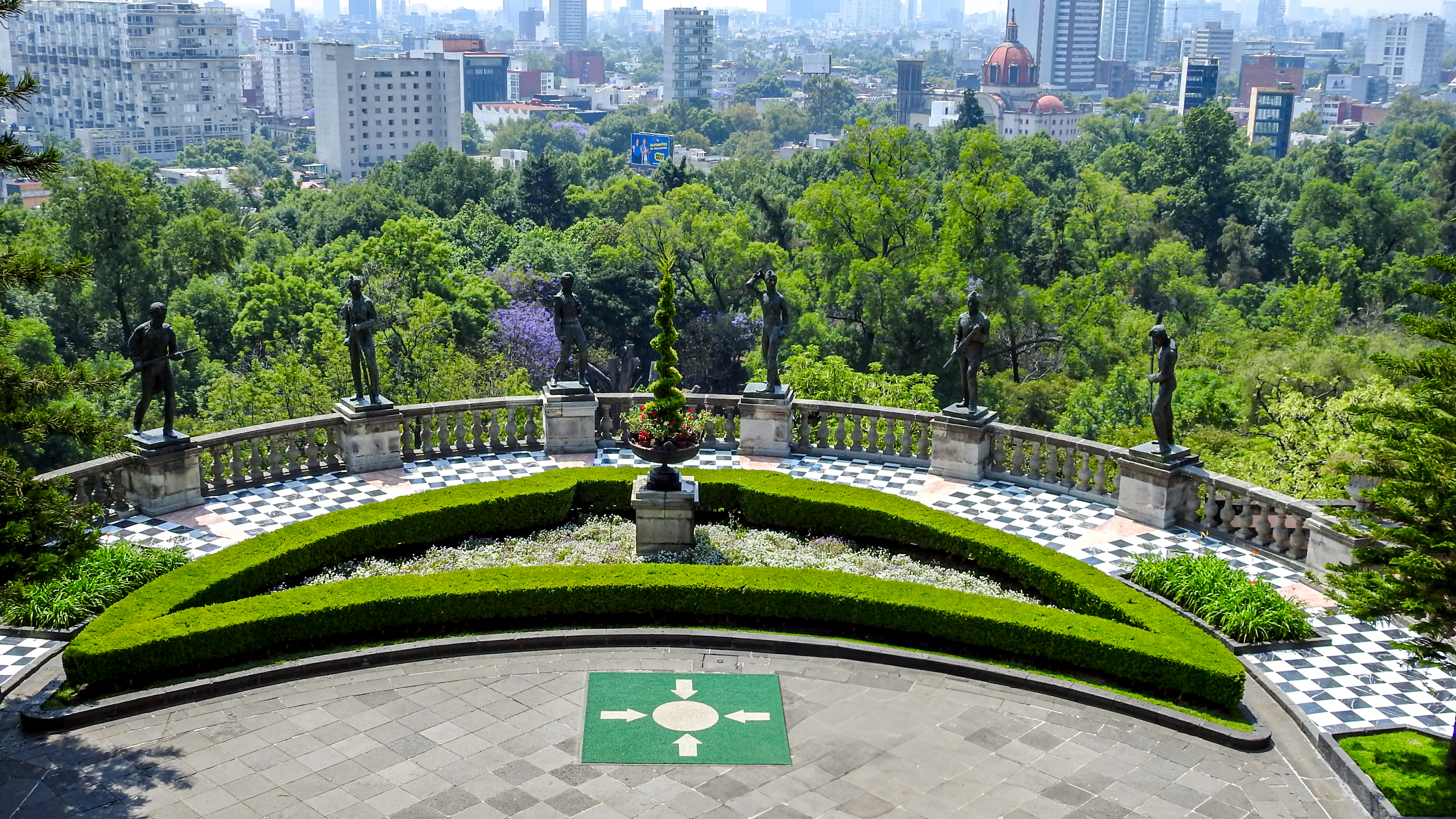 A panoramic view from Chapultepec Castle in Mexico City, overlooking a lush green park and the city skyline. The foreground features a manicured terrace with classical statues and a checkered tile floor. This breathtaking view is an inspiring backdrop for MICE industry events and incentive travel.