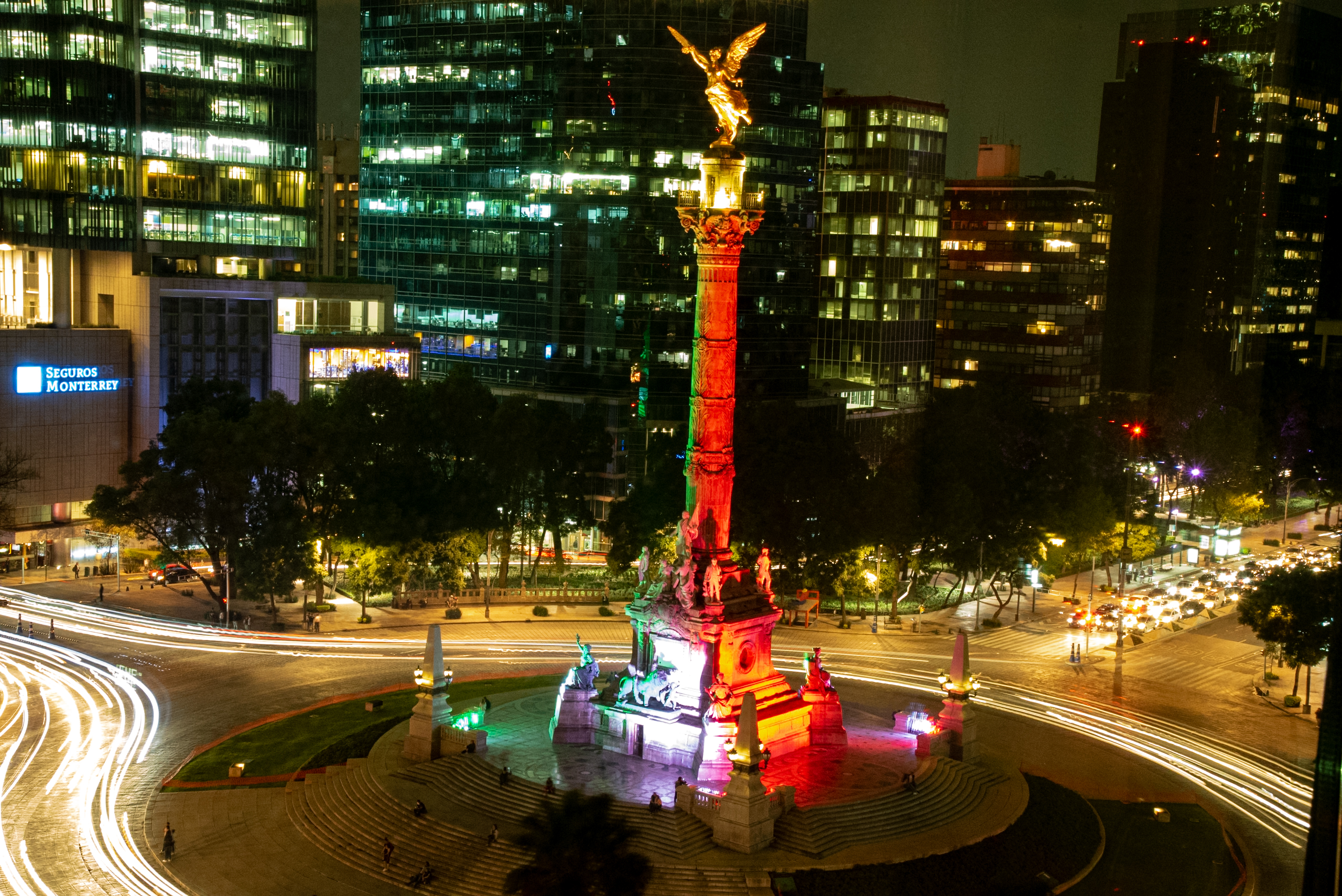 The Angel of Independence monument at night in Mexico City. This long-exposure photo shows streaks of light from traffic surrounding the monument, which is illuminated in red and green light. The monument is set on a roundabout with a grassy cantera stone base and is surrounded by modern, high-rise office buildings. The image highlights the city's vibrant urban atmosphere, making it an excellent destination for MICE industry events and incentive travel.